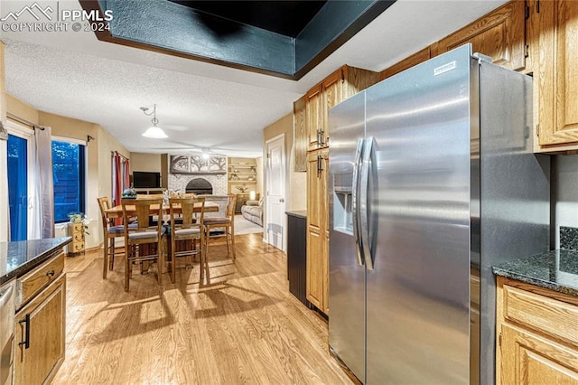 kitchen with a brick fireplace, stainless steel appliances, light wood-type flooring, a textured ceiling, and dark stone counters