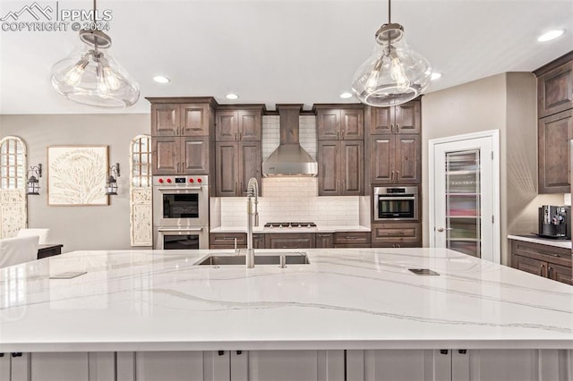 kitchen with a large island with sink, wall chimney range hood, hanging light fixtures, dark brown cabinetry, and appliances with stainless steel finishes