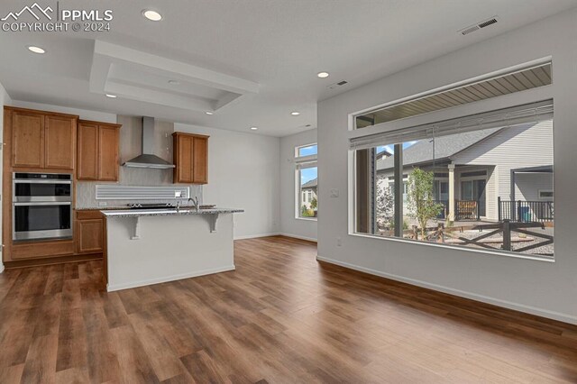 kitchen featuring an island with sink, wall chimney exhaust hood, dark wood-type flooring, stainless steel double oven, and a kitchen bar