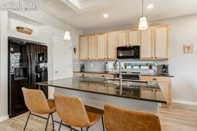 kitchen with light hardwood / wood-style floors, a kitchen island with sink, pendant lighting, dark stone counters, and black appliances