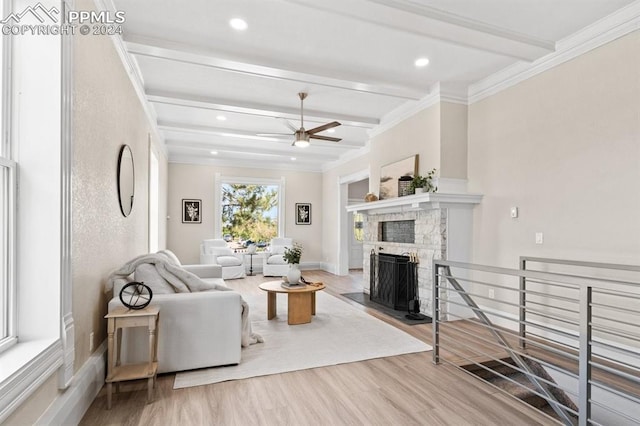 living room with a stone fireplace, light wood-type flooring, beam ceiling, ceiling fan, and ornamental molding