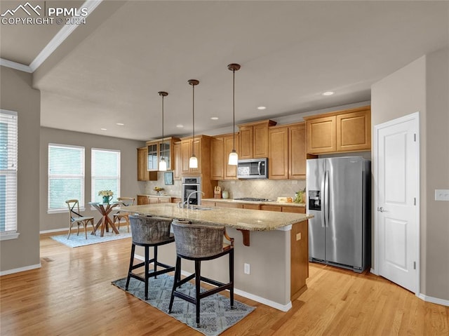 kitchen with appliances with stainless steel finishes, hanging light fixtures, light hardwood / wood-style floors, light stone countertops, and a center island with sink