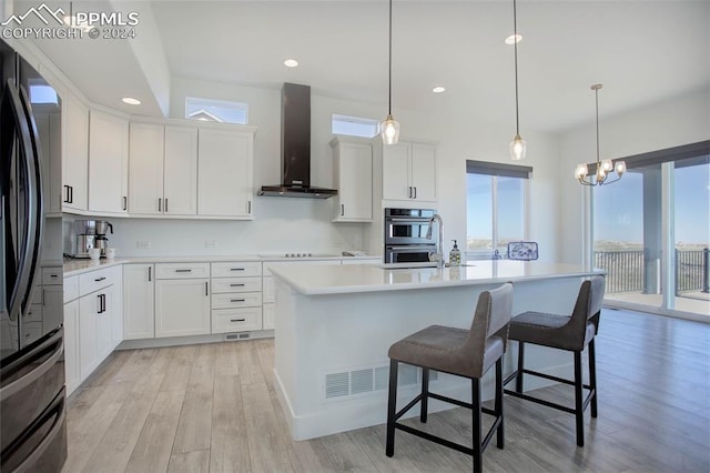 kitchen featuring pendant lighting, a kitchen island with sink, wall chimney range hood, white cabinetry, and light wood-type flooring