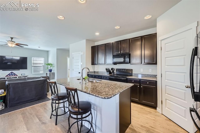 kitchen featuring an island with sink, light wood-type flooring, black appliances, dark stone counters, and sink