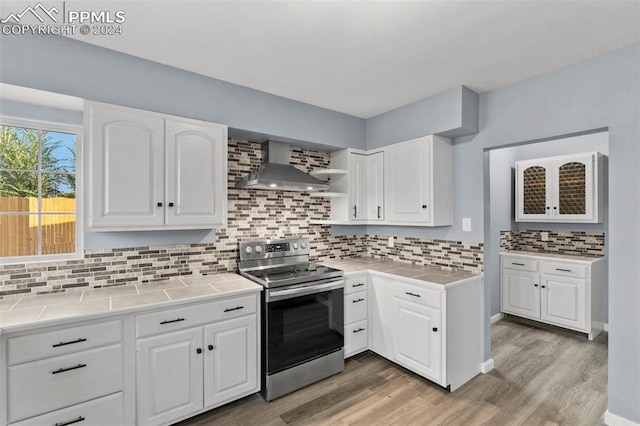 kitchen featuring wall chimney exhaust hood, tile counters, electric stove, and white cabinetry