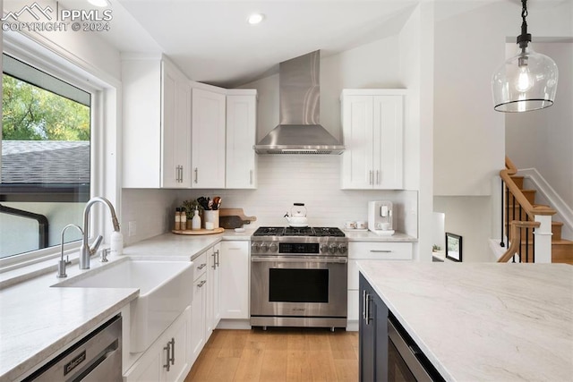 kitchen with hanging light fixtures, wall chimney exhaust hood, white cabinetry, stainless steel appliances, and light wood-type flooring