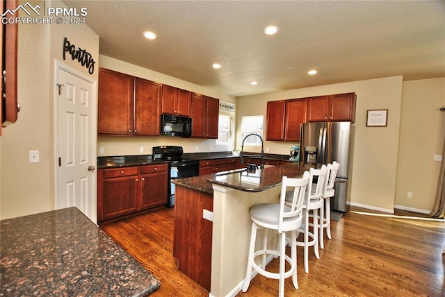 kitchen with dark stone counters, black appliances, dark hardwood / wood-style flooring, and a kitchen island with sink