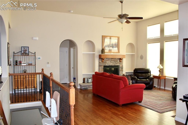 living room with arched walkways, wood finished floors, a high ceiling, a stone fireplace, and built in shelves