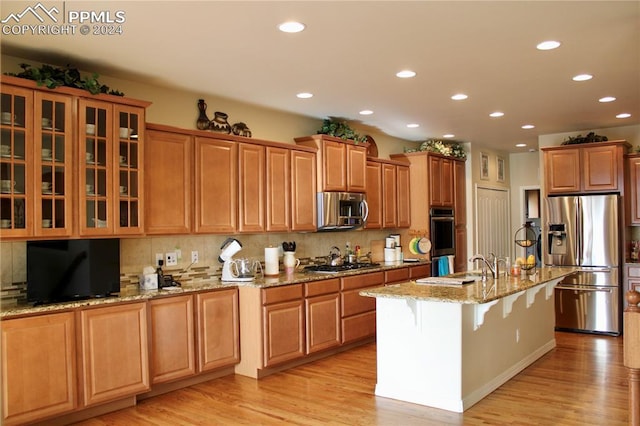 kitchen with a breakfast bar, a center island with sink, light stone countertops, appliances with stainless steel finishes, and light hardwood / wood-style floors