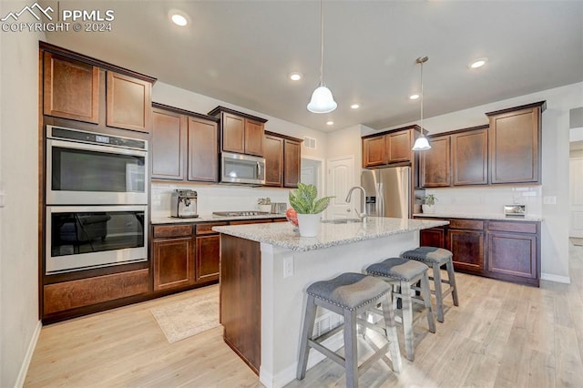 kitchen with appliances with stainless steel finishes, hanging light fixtures, light hardwood / wood-style floors, a breakfast bar area, and a kitchen island with sink