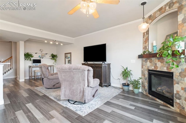 living room featuring ceiling fan, a stone fireplace, wood-type flooring, crown molding, and track lighting