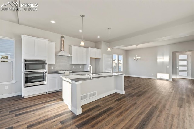 kitchen with white cabinetry, wall chimney exhaust hood, dark hardwood / wood-style floors, double oven, and an island with sink