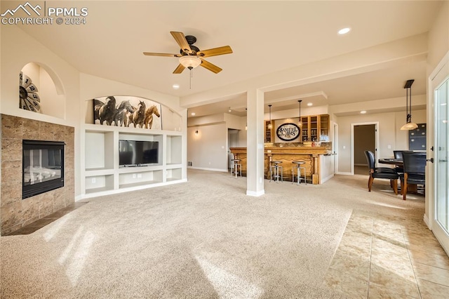 carpeted living room with bar area, a tiled fireplace, ceiling fan, and built in shelves