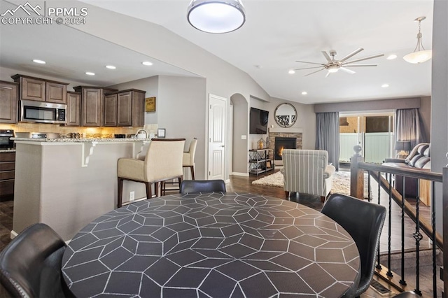 dining area with ceiling fan, a stone fireplace, dark wood-type flooring, and vaulted ceiling