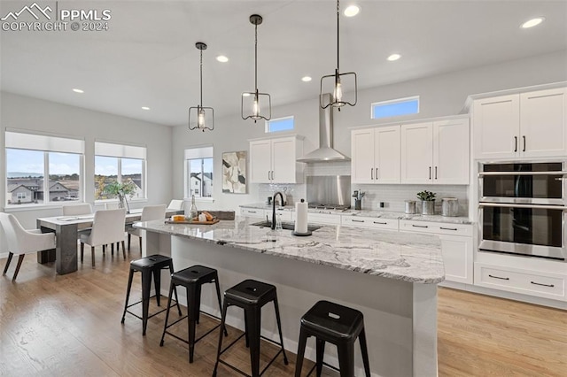 kitchen featuring light wood-type flooring, an island with sink, wall chimney range hood, and white cabinets