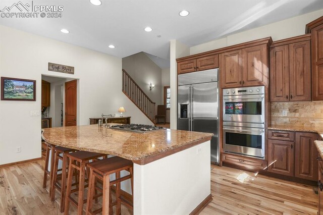 kitchen featuring a center island with sink, appliances with stainless steel finishes, light stone countertops, and light wood-type flooring