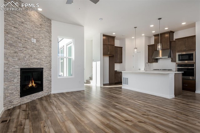 kitchen featuring appliances with stainless steel finishes, dark wood-type flooring, a center island with sink, a stone fireplace, and hanging light fixtures