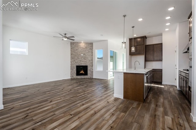 kitchen featuring a stone fireplace, decorative light fixtures, an island with sink, dark brown cabinets, and dark hardwood / wood-style flooring