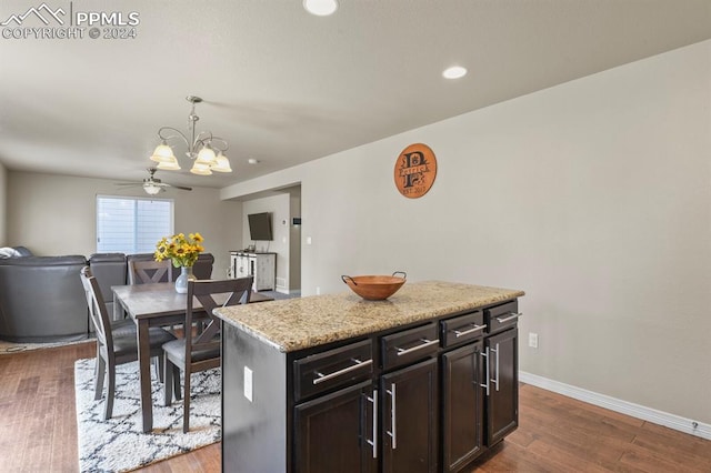 kitchen with a kitchen island, ceiling fan with notable chandelier, pendant lighting, dark wood-type flooring, and light stone counters