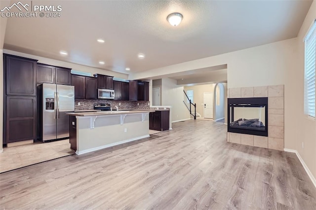 kitchen with a tiled fireplace, light hardwood / wood-style flooring, an island with sink, a breakfast bar, and appliances with stainless steel finishes