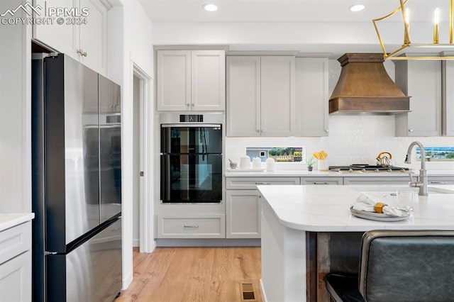 kitchen with decorative backsplash, decorative light fixtures, light wood-type flooring, custom exhaust hood, and appliances with stainless steel finishes