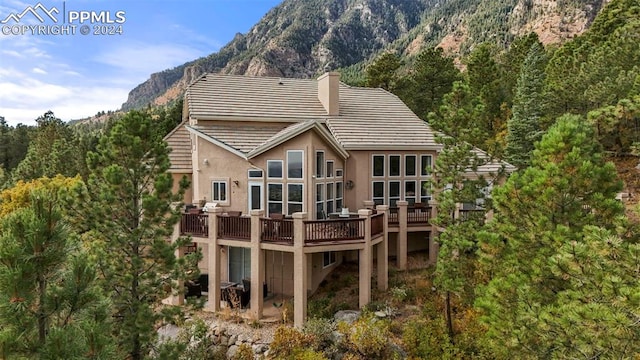 back of house featuring a deck with mountain view, stucco siding, a chimney, a view of trees, and a tile roof