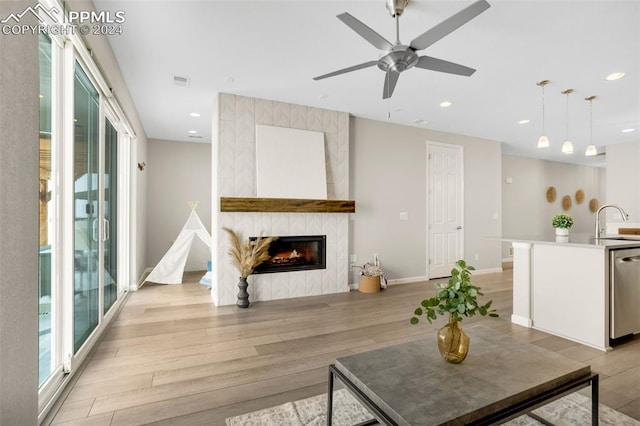 living room with ceiling fan, a tiled fireplace, sink, and light wood-type flooring