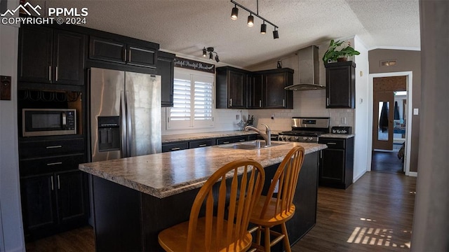 kitchen with lofted ceiling, wall chimney exhaust hood, an island with sink, sink, and appliances with stainless steel finishes