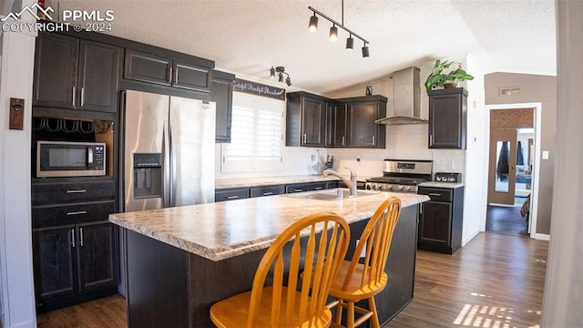 kitchen with wall chimney range hood, stainless steel appliances, a center island with sink, sink, and vaulted ceiling