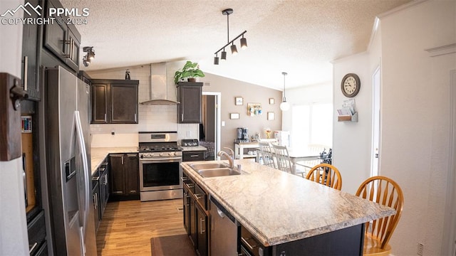 kitchen featuring appliances with stainless steel finishes, hanging light fixtures, wall chimney exhaust hood, vaulted ceiling, and a kitchen island with sink