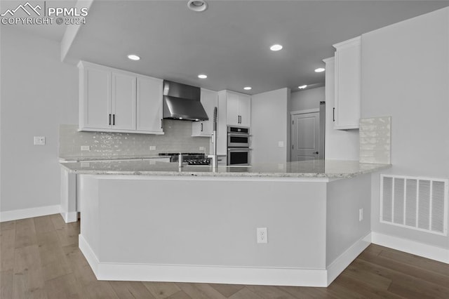 kitchen featuring white cabinets, wall chimney exhaust hood, kitchen peninsula, and double oven