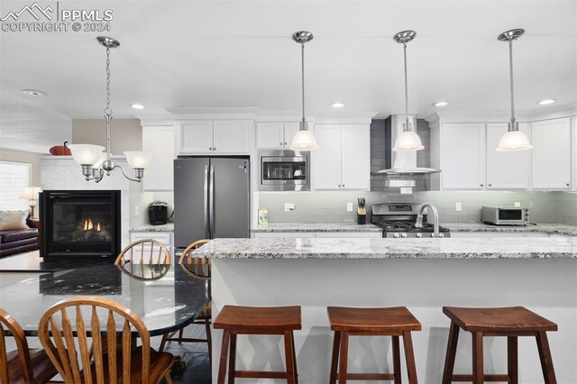 kitchen featuring stainless steel appliances, light stone counters, wall chimney exhaust hood, a tile fireplace, and pendant lighting