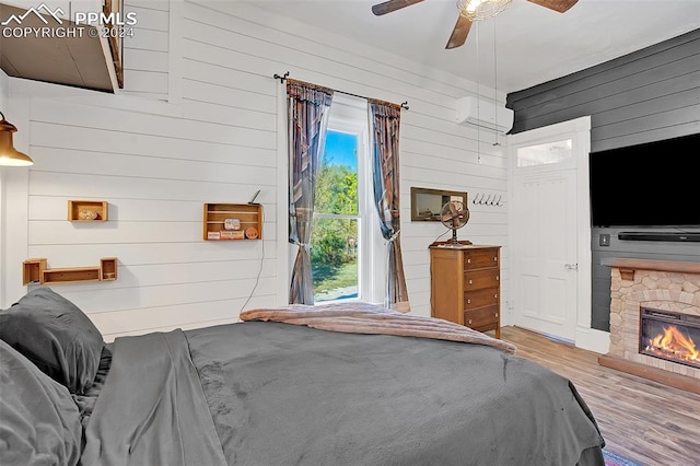 bedroom with wood-type flooring, a wall mounted AC, wood walls, ceiling fan, and a fireplace