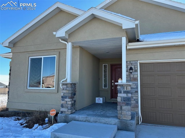 snow covered property entrance with a garage, stone siding, and stucco siding