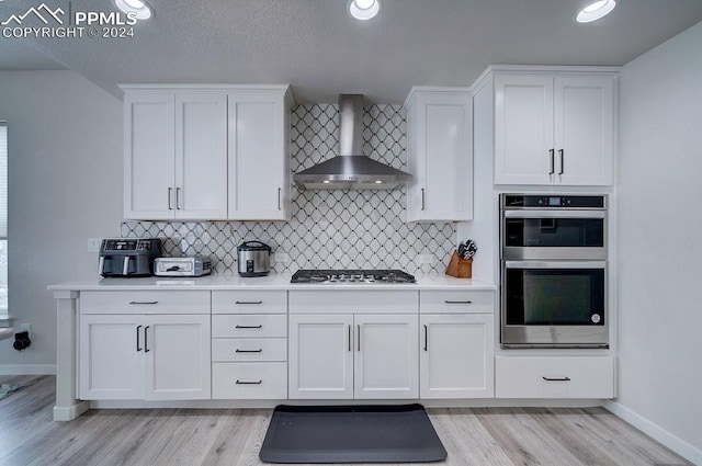 kitchen with wall chimney exhaust hood, stainless steel appliances, backsplash, light hardwood / wood-style floors, and white cabinets