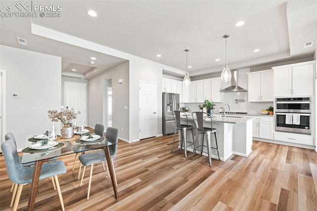 kitchen featuring appliances with stainless steel finishes, light wood-type flooring, a tray ceiling, wall chimney range hood, and white cabinetry