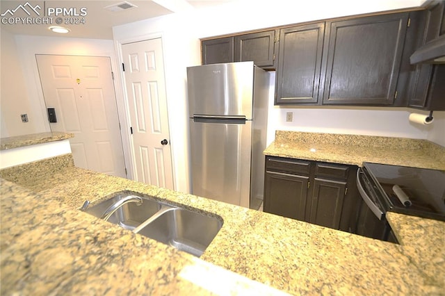 kitchen featuring light stone counters, dark brown cabinets, wall chimney range hood, sink, and stainless steel refrigerator