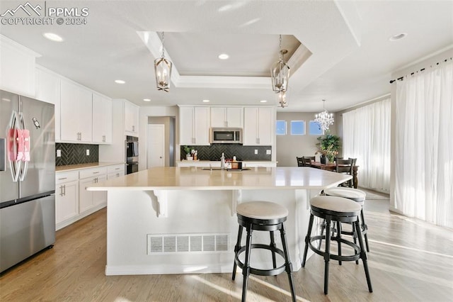 kitchen with appliances with stainless steel finishes, a tray ceiling, light hardwood / wood-style flooring, white cabinets, and an island with sink