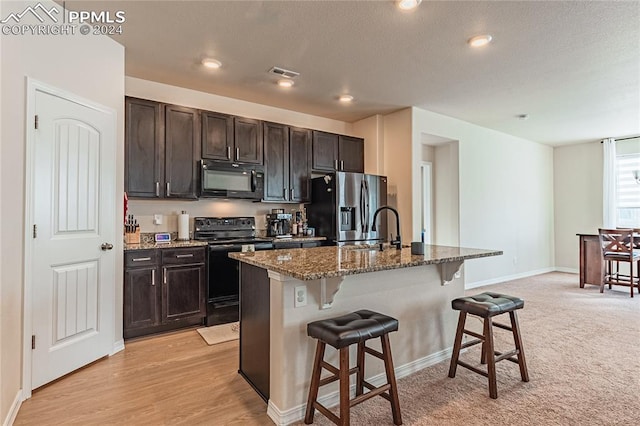 kitchen with black appliances, light hardwood / wood-style flooring, an island with sink, dark brown cabinets, and a breakfast bar area