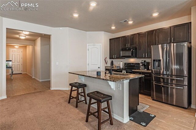 kitchen featuring dark stone counters, black appliances, a kitchen breakfast bar, a center island with sink, and light colored carpet