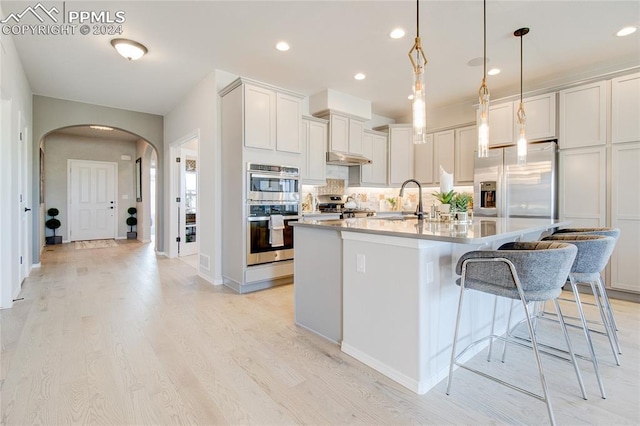 kitchen with pendant lighting, a kitchen island with sink, light wood-type flooring, appliances with stainless steel finishes, and tasteful backsplash