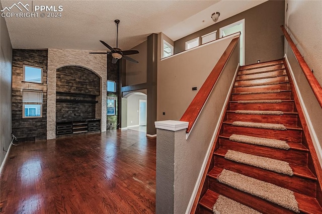 staircase with ceiling fan, high vaulted ceiling, wood-type flooring, a textured ceiling, and a fireplace