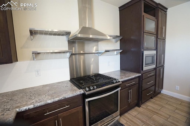 kitchen featuring appliances with stainless steel finishes, light stone counters, dark brown cabinets, wall chimney range hood, and light hardwood / wood-style floors