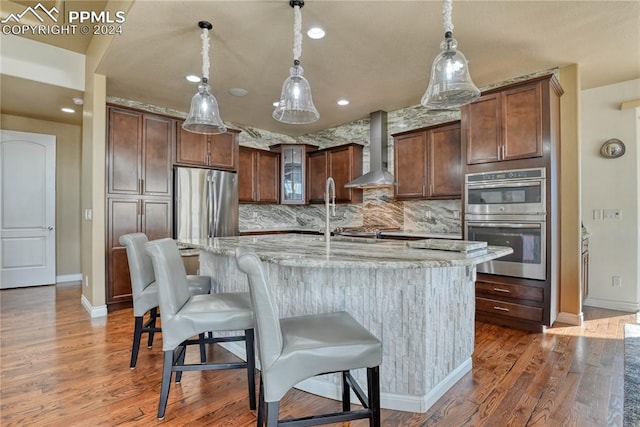 kitchen featuring dark wood-type flooring, wall chimney range hood, light stone counters, a kitchen island with sink, and appliances with stainless steel finishes