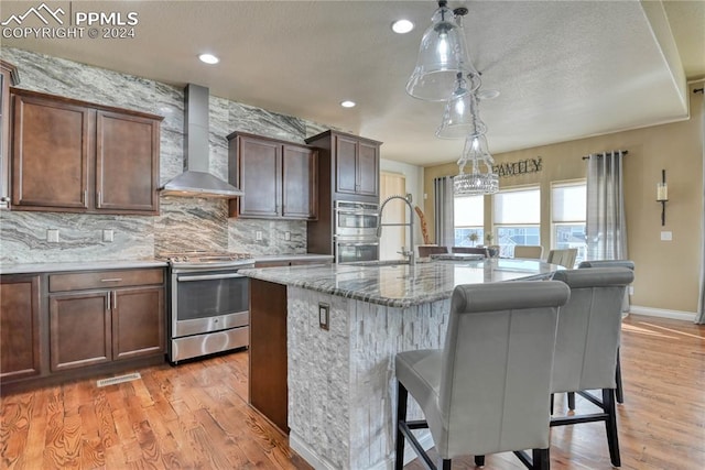 kitchen with light stone countertops, appliances with stainless steel finishes, wall chimney range hood, a center island with sink, and light hardwood / wood-style floors