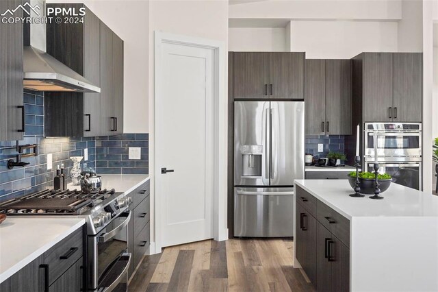 kitchen featuring appliances with stainless steel finishes, backsplash, wall chimney exhaust hood, dark brown cabinets, and wood-type flooring