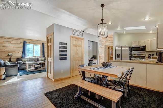 dining room featuring wooden walls, vaulted ceiling with skylight, a chandelier, and light wood-type flooring