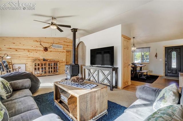 living room with vaulted ceiling, ceiling fan, wooden walls, wood-type flooring, and a wood stove