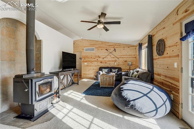 carpeted living room featuring ceiling fan, a wood stove, wooden walls, and vaulted ceiling