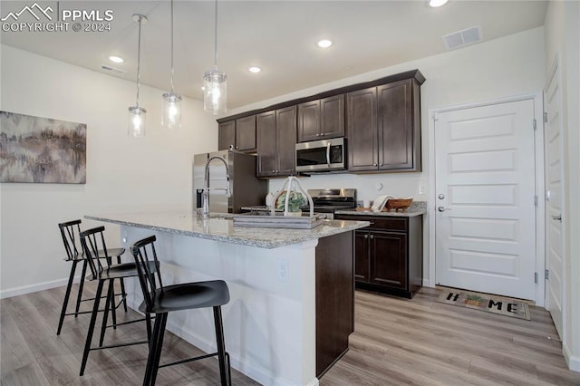 kitchen featuring a kitchen island with sink, hanging light fixtures, light wood-type flooring, appliances with stainless steel finishes, and dark brown cabinets